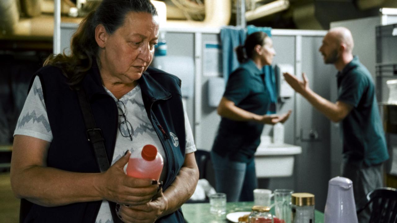 Still aus dem Drama ICH VERSTEHE IHREN UNMUT: Eine Frau, Ende 50, in Arbeirskleidung hält in einem Arbeitsraum eine Flasche mit Reinigungsmittel in den Händen. Im Hintergrund streiten sich ein Mann und eine Frau.