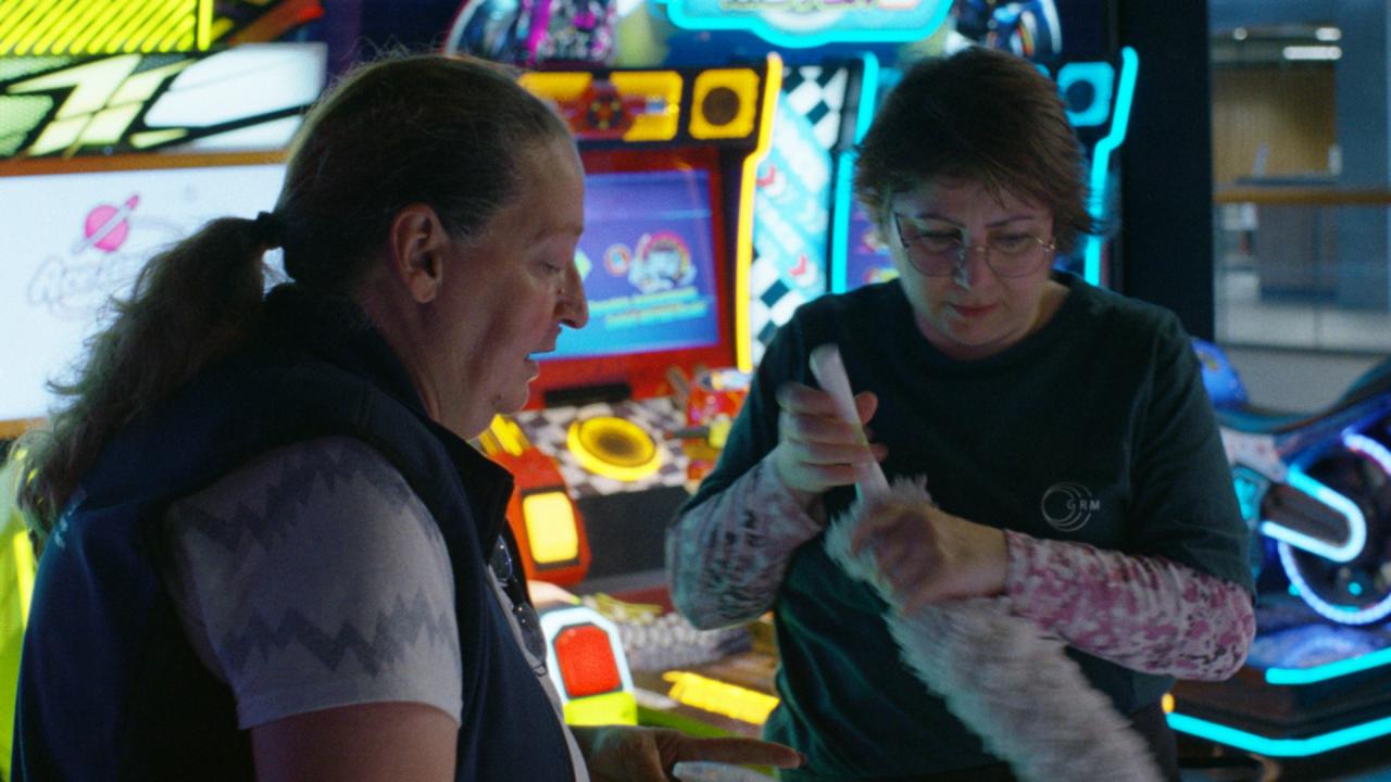 Still aus dem Film ICH VERSTEHE IHREN UNMUT: Halbnahe Aufnahme von zwei Frauen vor bunten Spielautomaten. Die rechte von beiden hält einen Staubwedel in den Händen.
