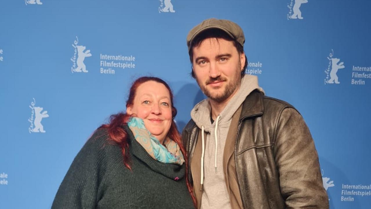 Halbnahe Aufnahme der Darstellerin Sabine Thalau und des Regisseurs Kilian Armando Friedrich: Beiden stehen nebeneinder vor einer blauen Wand mit der Aufschrift "Internationale Filmfestspiele Berlin" und blicken in die Kamera.