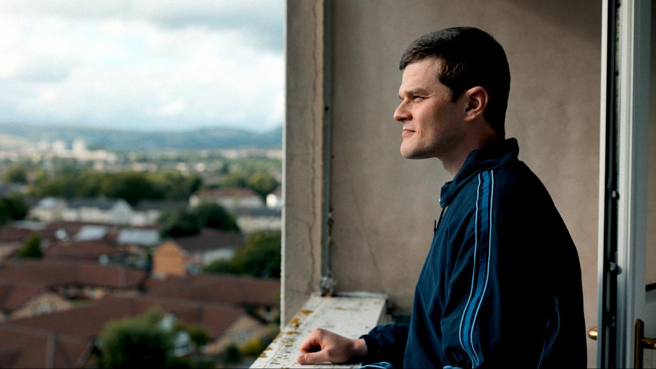 Still aus dem Drama VERFLUCHT NORMAL: Ein junger Mann in blauer Trainingsjacke steht auf einem Balkon und schaut auf eine Stadt. Er ist im Profil zu sehen.