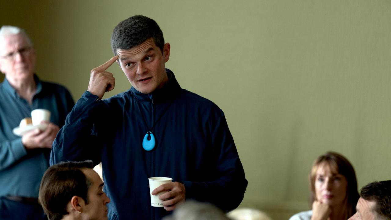 Still aus dem Drama VERFLUCHT NORMAL: Ein junger Mann steht in einer Gruppe von Meschen, die ihm zuhören. Er zeigt mit seinem rechten Zeigefinger an seine Stirn.