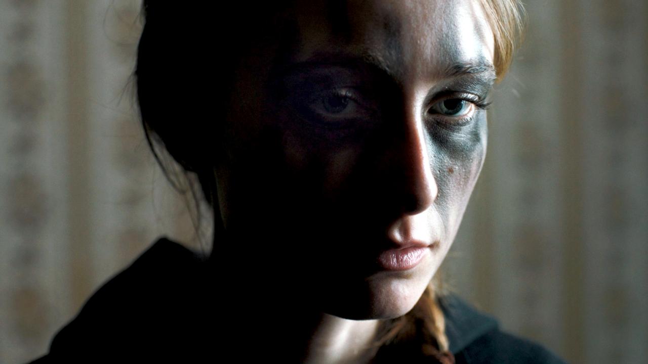 Still aus dem Drama WOLVES: Großaufnahme einer jungen Frau, deren Augenpartie schwarz geschminkt ist. Ihr Gesicht liegt zur Hälfte im Dunkeln. Sie schaut ernst direkt in die Kamera.