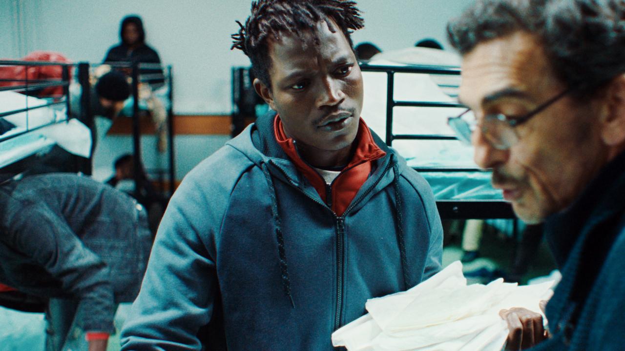 Still aus dem Film SOULEYMANS GESCHICHTE: Im Mittelpunkt des Halbnahaufnahme sit ein Mann zu sehen, der Bettbeüge im Arm hält und zu einem anderen Mann mit Briile rechts im Vordergrund blickt. Dieser ist leicht unscharf. Im Hintergrund sind ebenfalls unscharf weitere Personen und mehrere Etagenbetten zu erkennen.