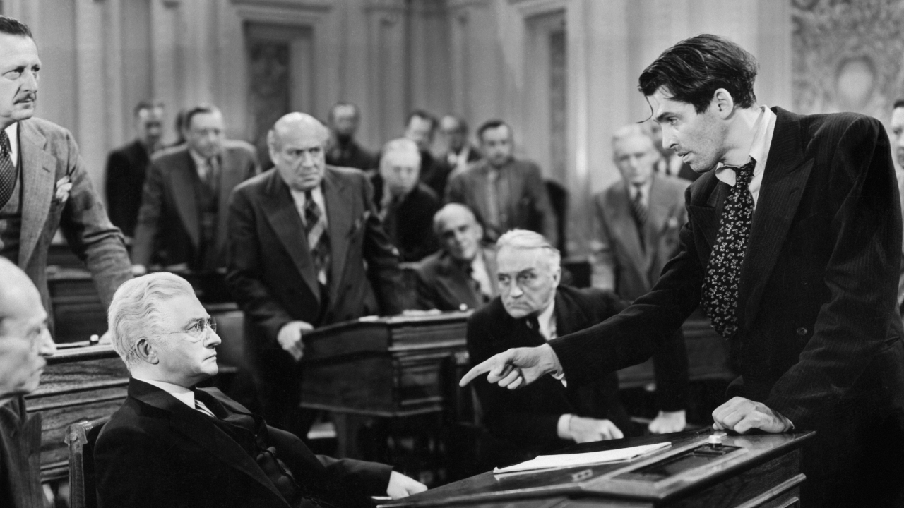 Filmstill aus dem Drama MR. SMITH GEHT NACH WASHINGTON: Schwarzweißaufnahme eines Plenarsaals, in dem ein stehender Mann mit ausgestrecktem Zeigefinge auf einen vor ihm hinter einem Tisch sitzenden Mann weist und offenbar erregt auf ihn einredet. Die Auseinandersetzung wird von zahlreichen ernst blickenden Männern beobachtet.