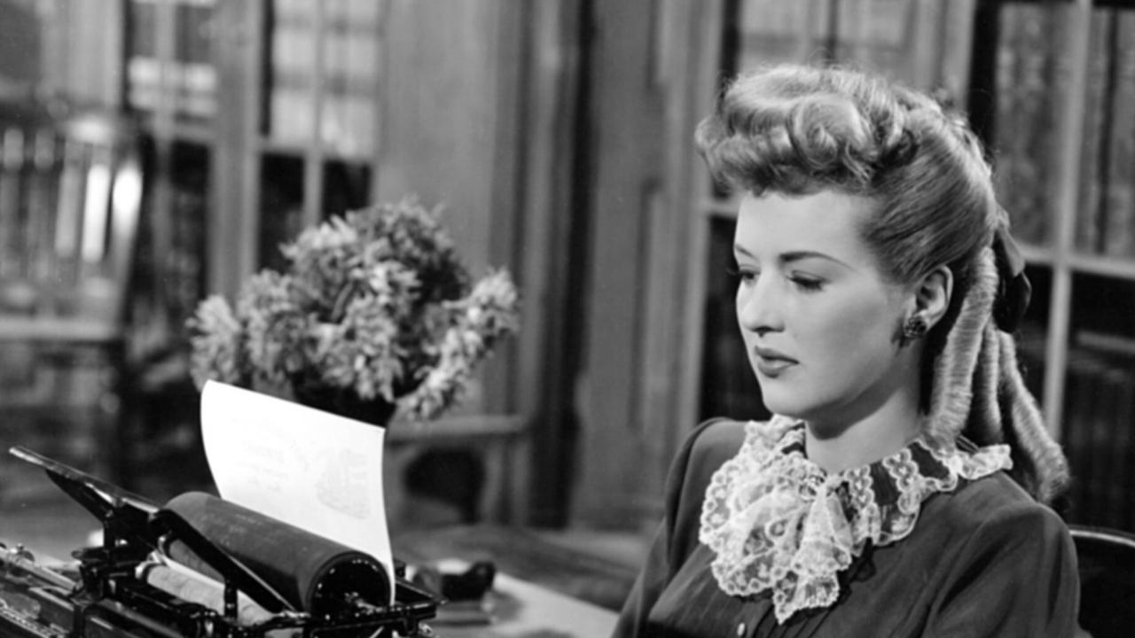 Still aus dem Film THE SHOCKING MISS PILGRIM: Eine junge Frau, gekleidet im Stil des 19. Jahrhunderts, sitzt an einer Schreibmaschine und tippt.