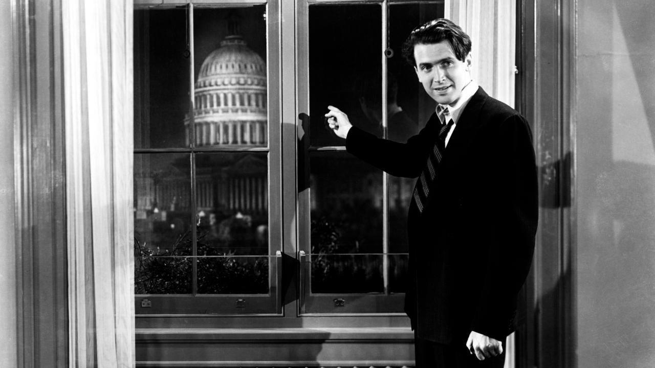 Still aus dem Drama MR. SMITH GEHT NACH WASHINGTON: Die Titelfigur steht an einem Fesnter und zeigt auf das erleuchtete Capitol, das im Hintergrund zu erkennen ist.