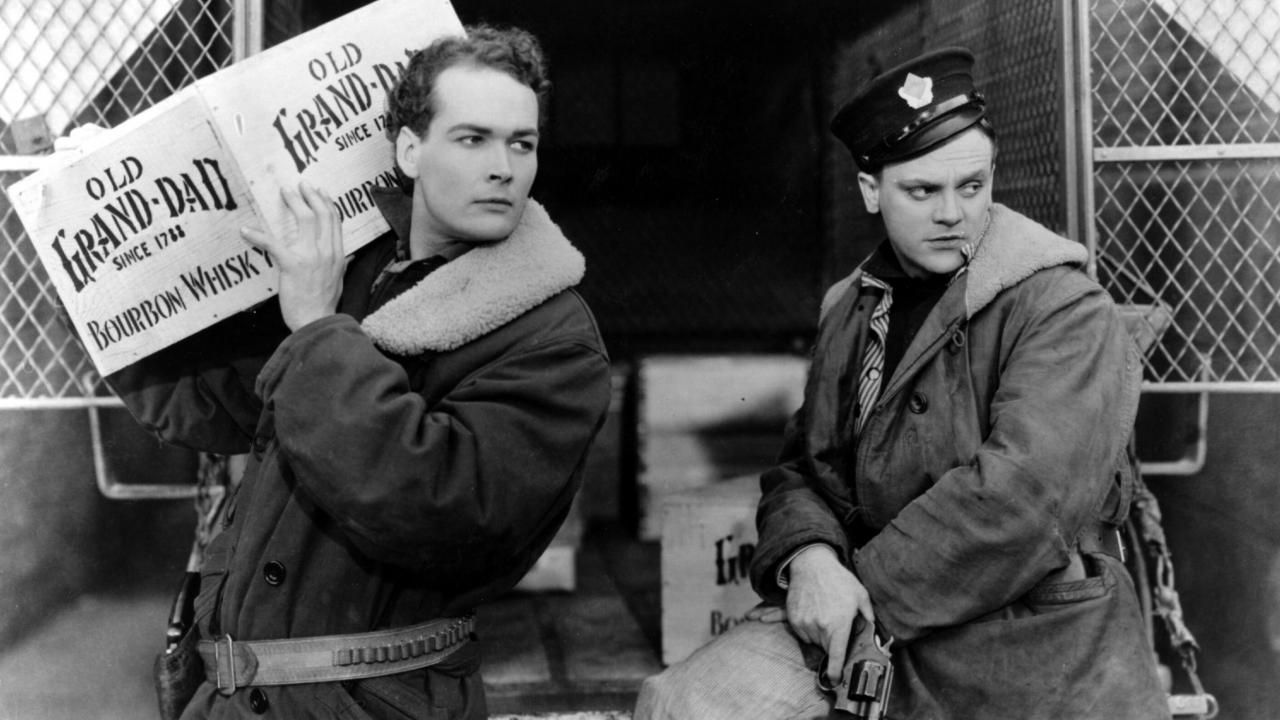 Still aus dem Gangsterfilm DER ÖFFENTLICHE FEIND (1931): Zwei leger gekleidete Männer schauen beide in die gleiche Richtung. Der eineträgt eine Kiste mit Whisky auf den Schultern, der andere hält eine Pistole in der Hand.