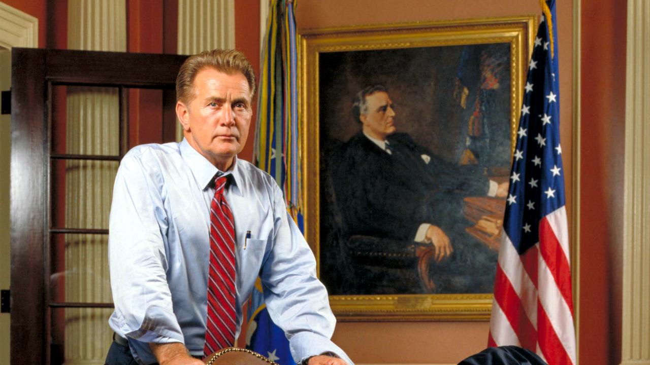 Standbild für die Serie THE WEST WING – IM ZENTRUM DER MACHT: Martim Sheen in der Rolle des US-Präsidenten, der in einem Büro steht. Neben ihm hängt die US-Flagge.