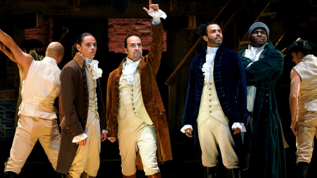 Still aus der Musical-Verfilmung HAMILTON: Vier im Stil des 18. Jahhunderts gekleidet Männer stehen nebeneinander, Die Figur des Hamilton streckt einen Arm in die Höhe.