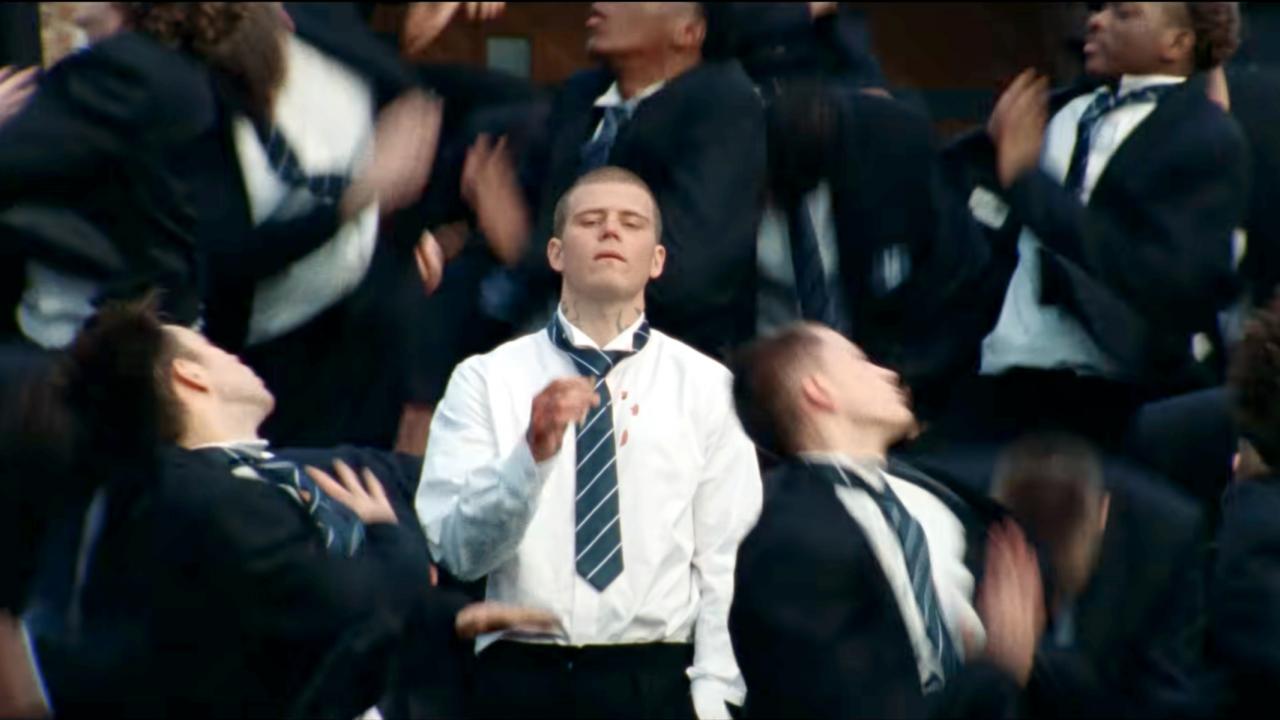 Screenshot aus dem Musikclip GENER8ION & YUNG LEAN: STORM: Ein junger Mann im weißen Hemd und mit loser Krawatte zwischen vGleichaltrigen, die sich hin und her bewegen. Sie tragen alle dunkle Anzüge.