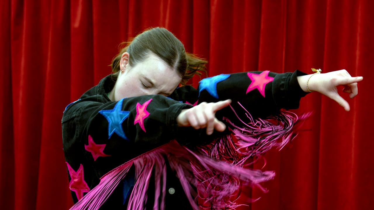 Still aus dem Drama SUNNY DANCER: Eine Teenagerin steht vor einem roten Vorhand und tanzt expressiv mit geschlossenen Augen. Sie trägt eine schwarze Jacke mit langen Fransen und bunten aufgenähten Sternen.