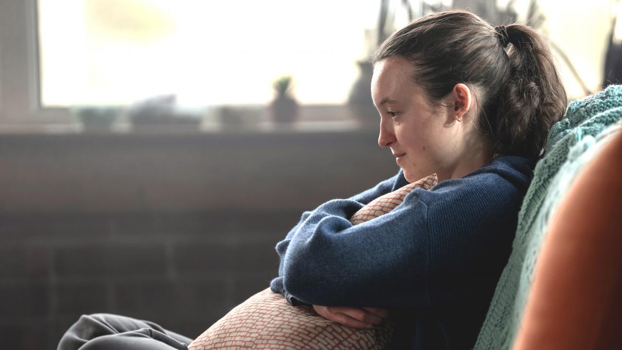 Still aus dem Drama SUNNY DANCER: Eine Teenagerin mit Pferdeschwanz sitzt auf einem Sofa und hält ein Kissen vor ihren Bauch. Sie ist im Profil zu sehen.