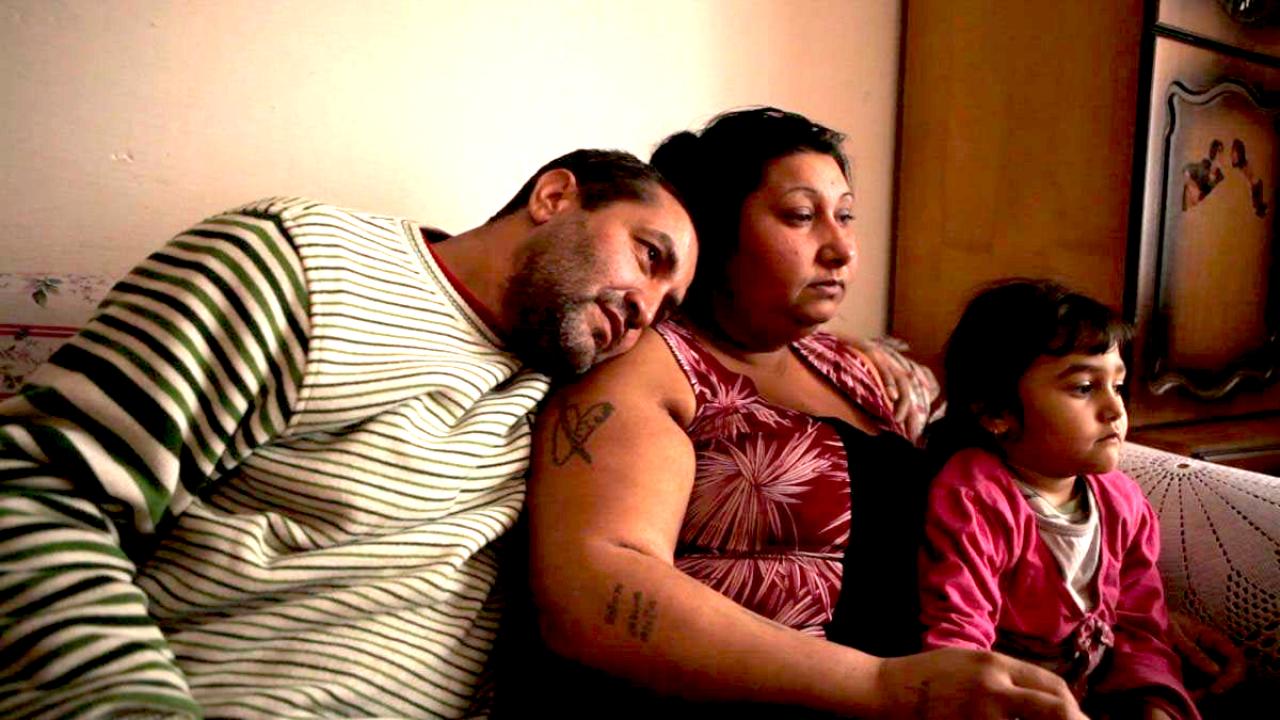Still aus dem Dokumentarfilm: AUS DEM LEBEN EINES SCHROTTSAMMLERS: Familie sitzt zusammen auf einem Sofa. Der Mann lehnt seinen Kopf auf die Schulter der Frau, das Kind sitzt neben ihnen.
