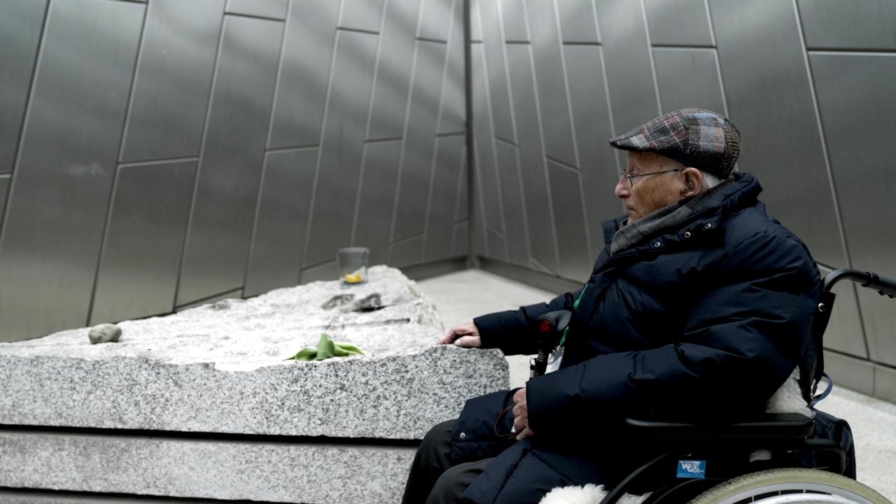 Still aus dem Dokumentarfilm ALBRECHT WEINBERG – ES IST IMMER IN MEINEM KOPF: Albrecht Weinberg sitzt in der Gedenkstätte Bergen-Belsen in seinem Rollstuhl und legt eine Hand auf einen Gedenkstein.