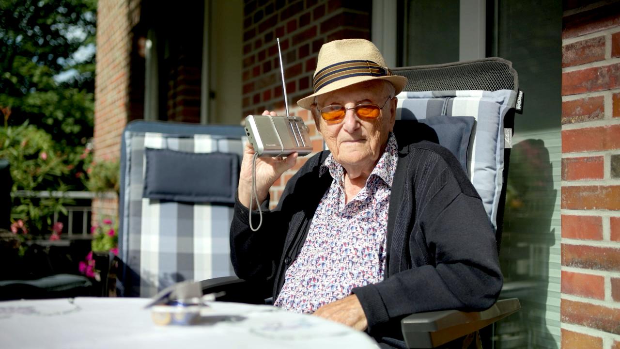 Still aus dem Dokumentarfilm ALBRECHT WEINBERG – ES IST IMMER IN MEINEM KOPF: Albrecht Weinberg, hoch betagt, sitzt auf einer Terrasse und hät ein kleines Radiogerät an sein Ohr.