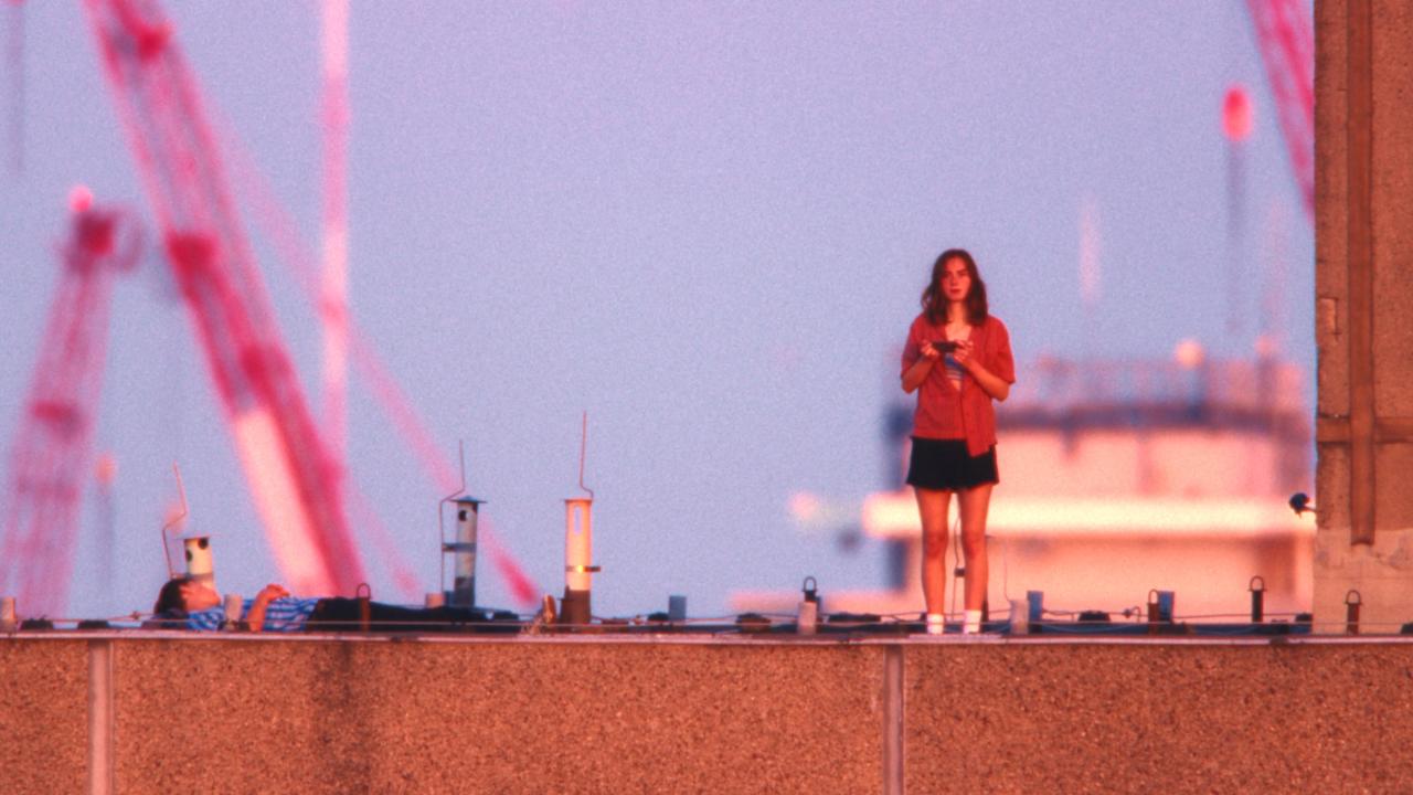 Still aus dem Drama EVERYTIME: Auf einem Hochaus steht eine Teenagerin, neben ihr liegt ein etwa gleichaltriger Junge. Im Hintergrund sind rote Kräne und ein anderes Hochausdach zu erkennen. Der Himmel ist leicht violett.