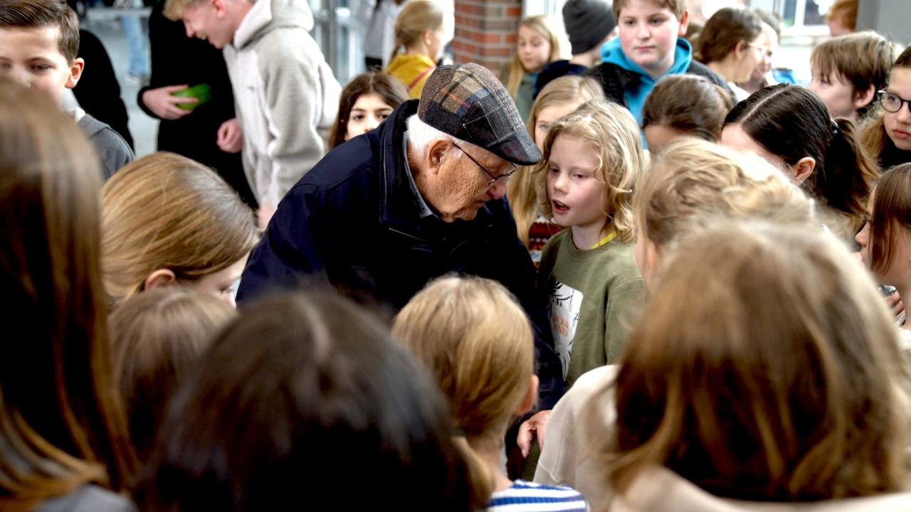 Still aus dem Dokumentarfilm ALBRECHT WEINBERG: Ein alter Mann mit Schirmmütze steht inmitten einer Gruppe von Schüler/-innen.