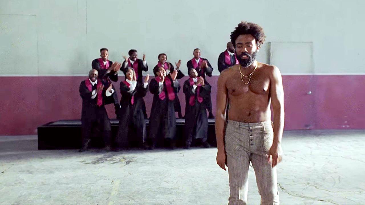 Screenshot aus dem Musikclip THIS IS AMERICA: Der Sänger Childish Gambino steht mit nacktem Oberkörper vor einem singenden Gospelchor, der vor einer Wand steht.