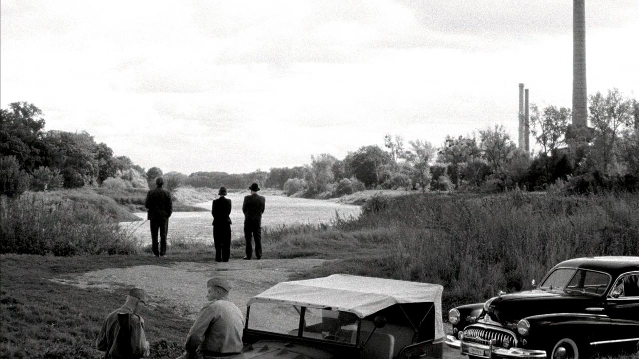 Still aus dem Drama VATERLAND: Ein Schwarz-Weiß-Bild: Zwei Männer und eine Frau stehen mit dem Rücken zur Kamera an einem Fluss und schauen in die Landschaft. Rechts sind Industrie-Schornsteine zu erkennen. Im Vordergrund steht eine alte Limousine und ein Militärjeep mit zwei Soldaten.