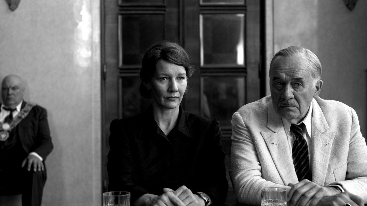 Still aus dem Drama VATERLAND: Eine Frau und ein älterer Mann sitzen nebeneinander an einem Tisch und scheinen jemanden zuzuhören. Es sollen Erika und Thomas Mann sein. Dsa Bild ist in Schwarz-Weiß.