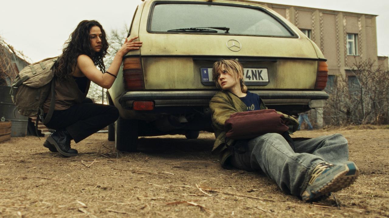 Szenenbild aus der Miniserie CITY OF BLOOD: Auf staubigem ausgetrockneten Boden lehnt sich eine liegende junge Frau  an die Stoßstange eines alten Mercedes-Kombis, während eine weitere junge Frau links neben dem Auto hockt. Im Hintergrund sind graue Gebäude und vertrocknete Sträucher zu sehen.