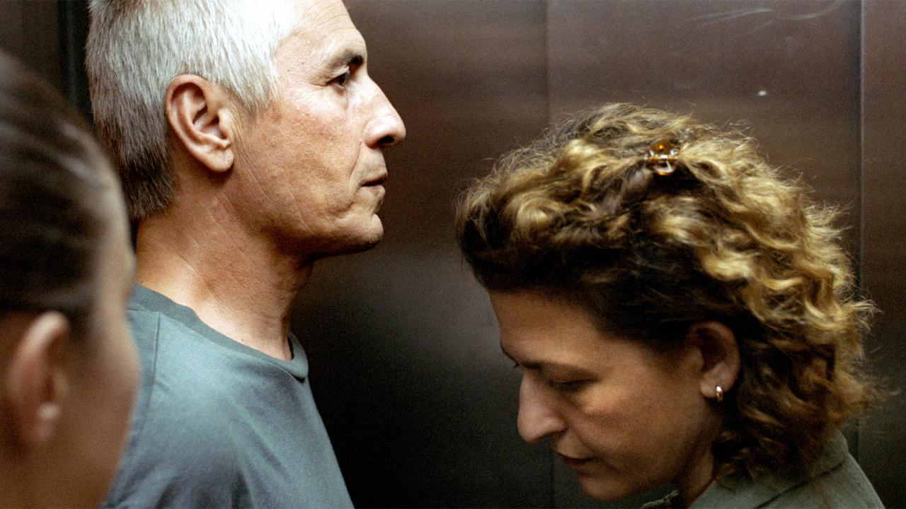 Still aus dem Drama VON SCHAM UND GELD: Ein Mann und eine Frau mittleren Alters stehen sich in einem beengtem Raum gegenüber. Sie sehen ernst oder besorgt aus. Sie sind im Profil zu sehen.
