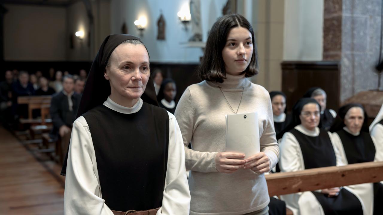 Still aus dem Drama GLAUBE: Eine Teenagerin in weißem Rollkragenpulli steht in einem Kirchengang neben einer Nonne. Auf den Bänken der Kirche sitzen weitere Nonnen.