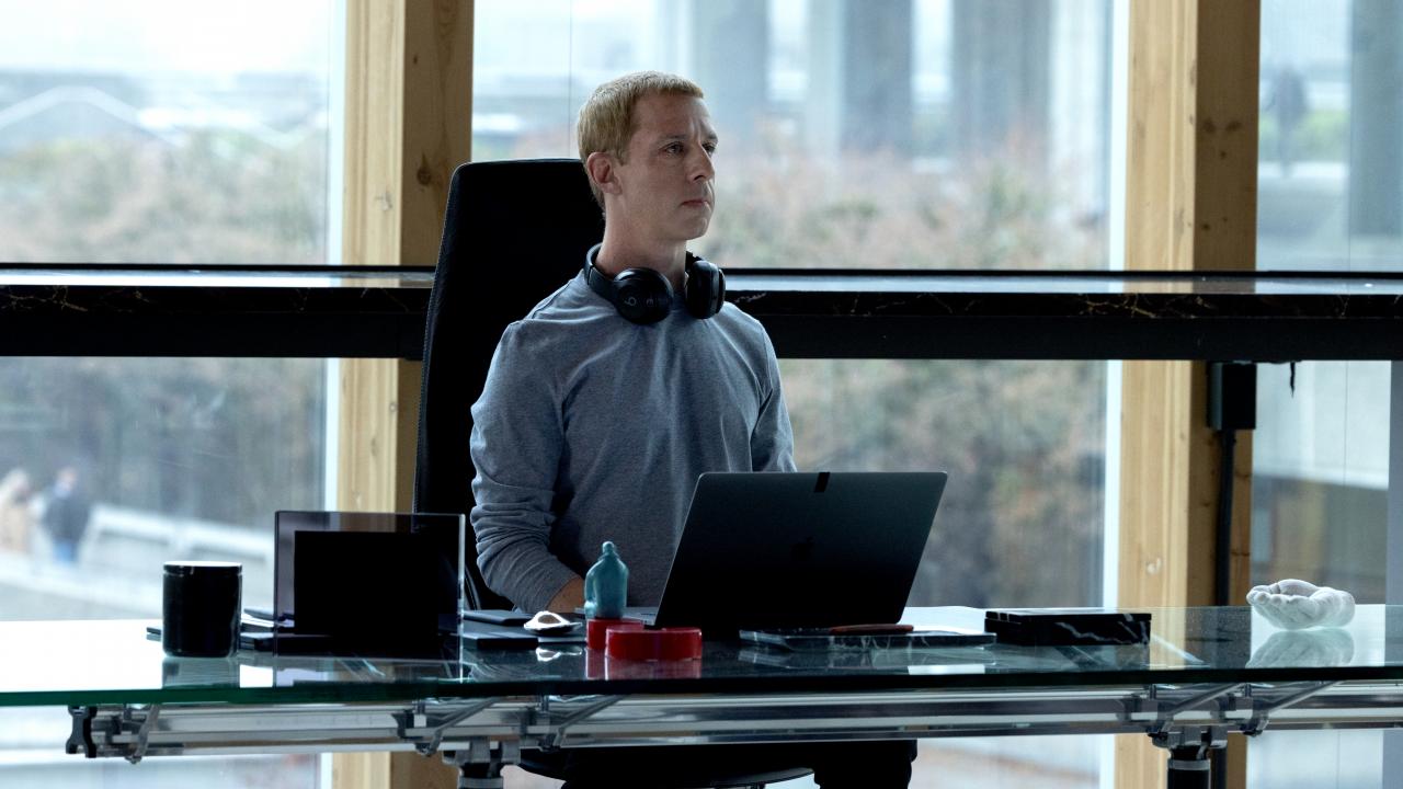 Still aus dem Drama THE SOCIAL RECKONING: Ein Mann, es soll Mark Zuckerberg sein, sitzt vor einer Fesnterwand an einem Schreibtisch. Er trägt ein Sweatshirt und Kopfhörer um den Hals. Er sitzt sehr gerade und schaut geradeaus.