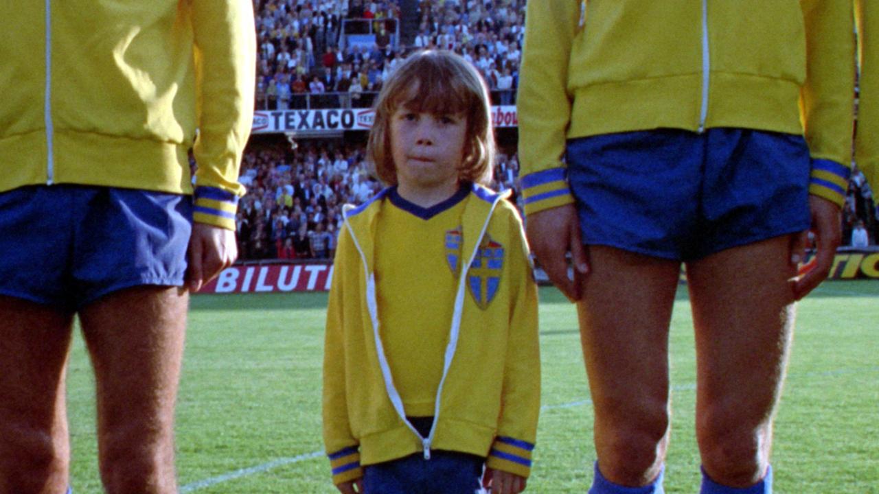 Fimpen, der Knirps,Szenenbild: Ein kleiner Junge im Trikot der schwedischen Nationalmannschaft steht neben seinen erwachsenen Teamkameraden, die aber nur bis zur Brust zu sehen sind.