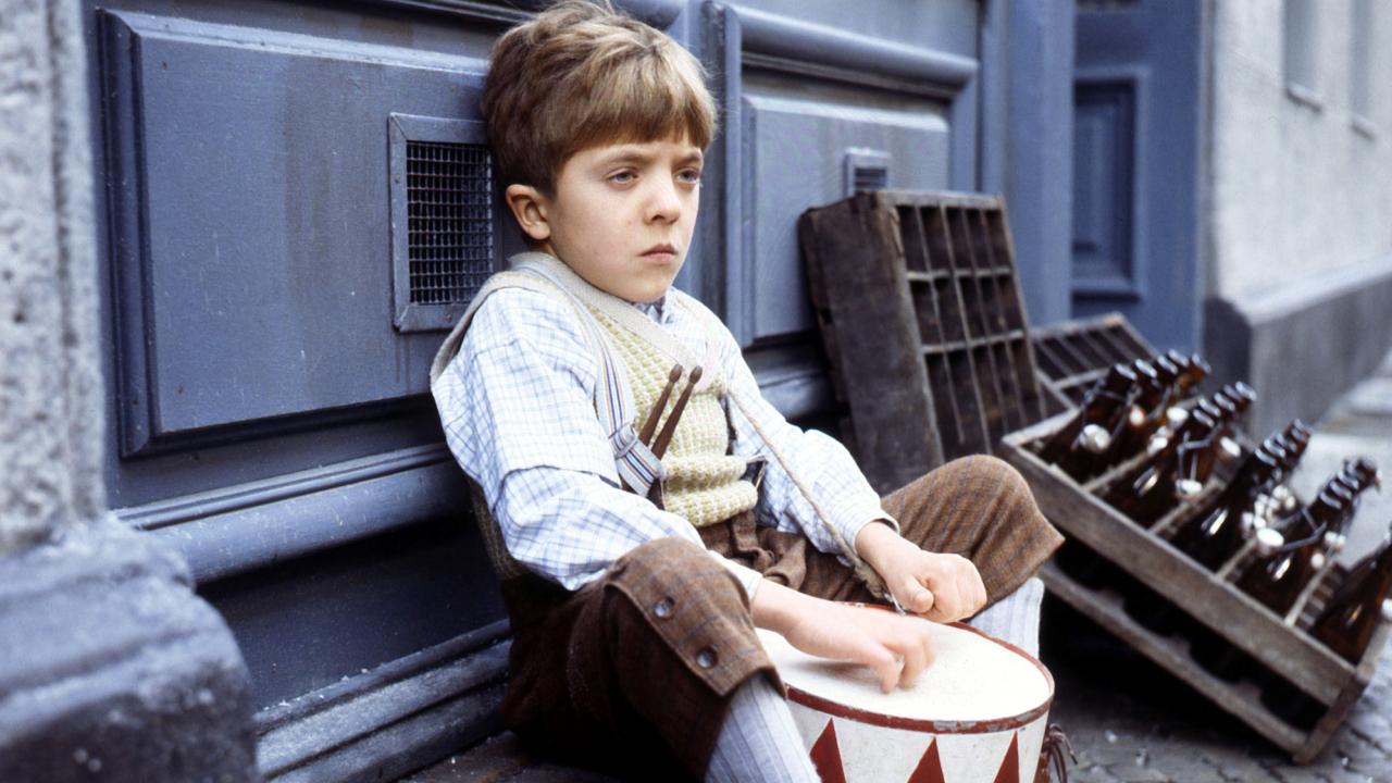 Filmstill aus dem Film DIE BLECHTROMMEL: Nahaufnahme eines blonden Jungen, der auf dem Boden sitzt und eine Blechtrommel im Schoß liegen hat.