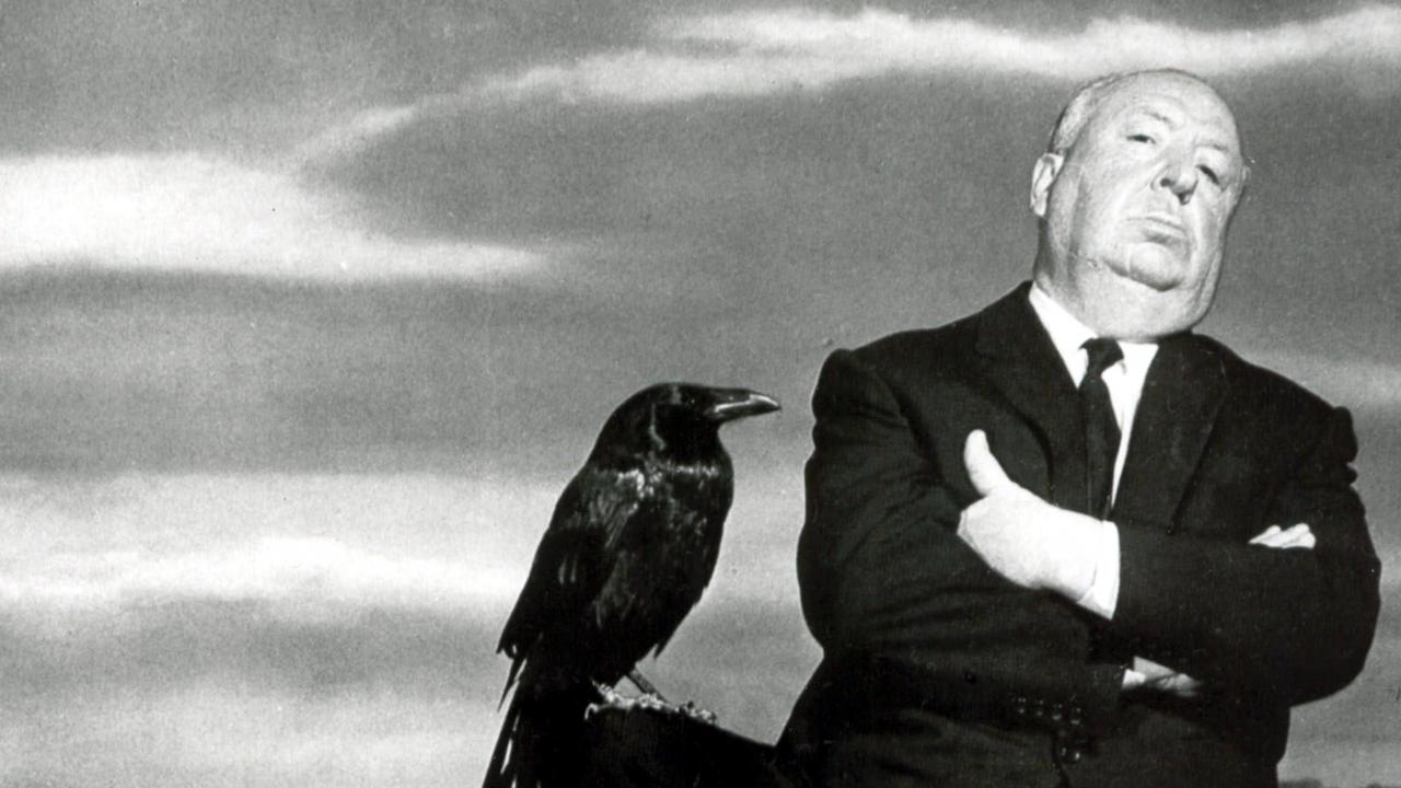 Publicityfoto: Halbnahaufnahme des Regisseurs Alfred Hitchcock: Er steht mit verschränkten Armen vor einem wolkigen Himmel und schaut herausfordernd in die Kamera. Auf seinem rechten, angewickelten Knie sitzt ein großer schwarzer Vogel.