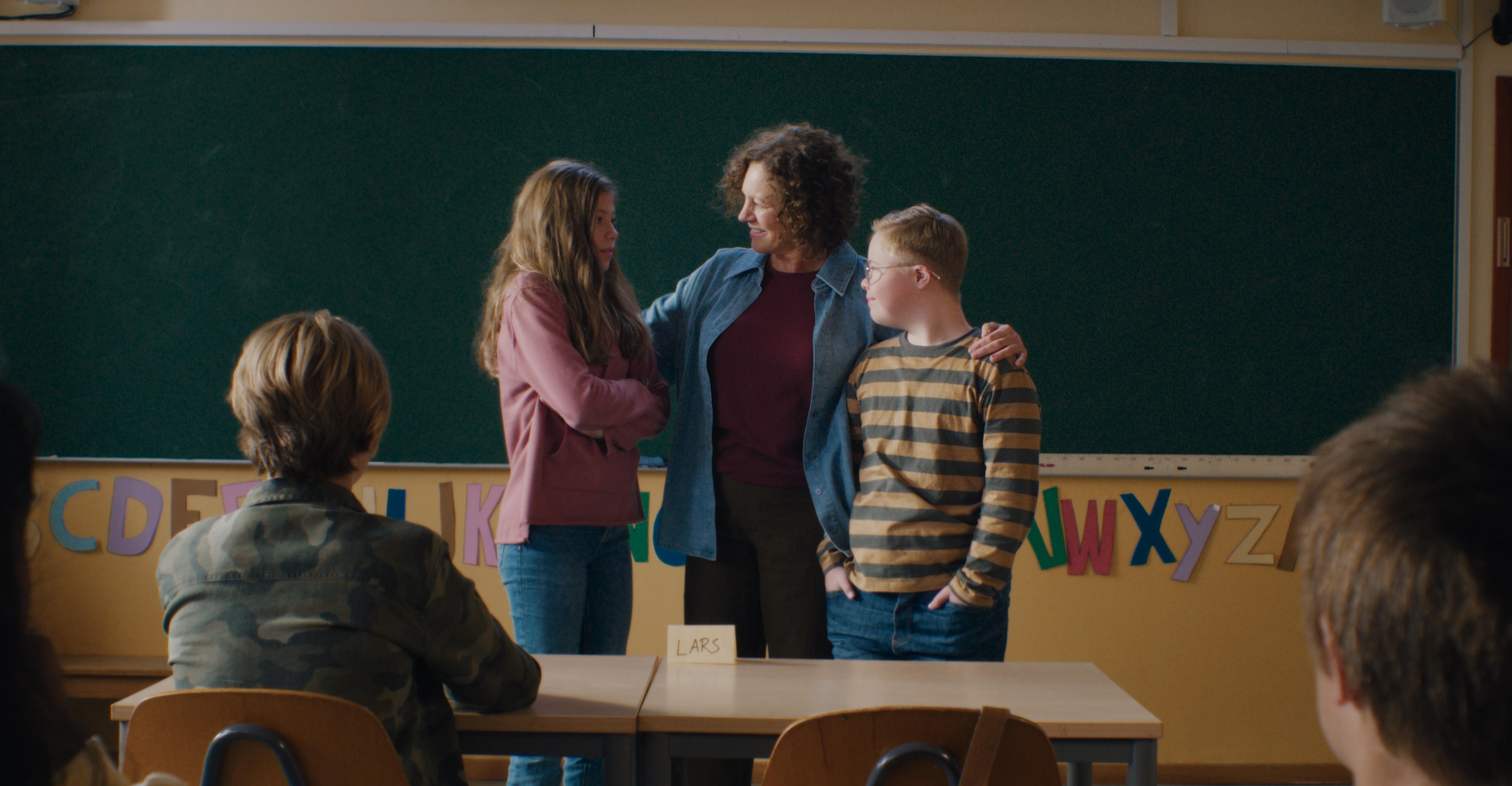 Filmstill aus LARS IST LOL: Vor einer Schultafel steht eine Frau, die ein Mädchen und einen Jungen miteinander bekannt macht. Das Mädchen hat die Arme verschränkt, der Junge lächelt sie an. Er hat das Down-Syndrom.