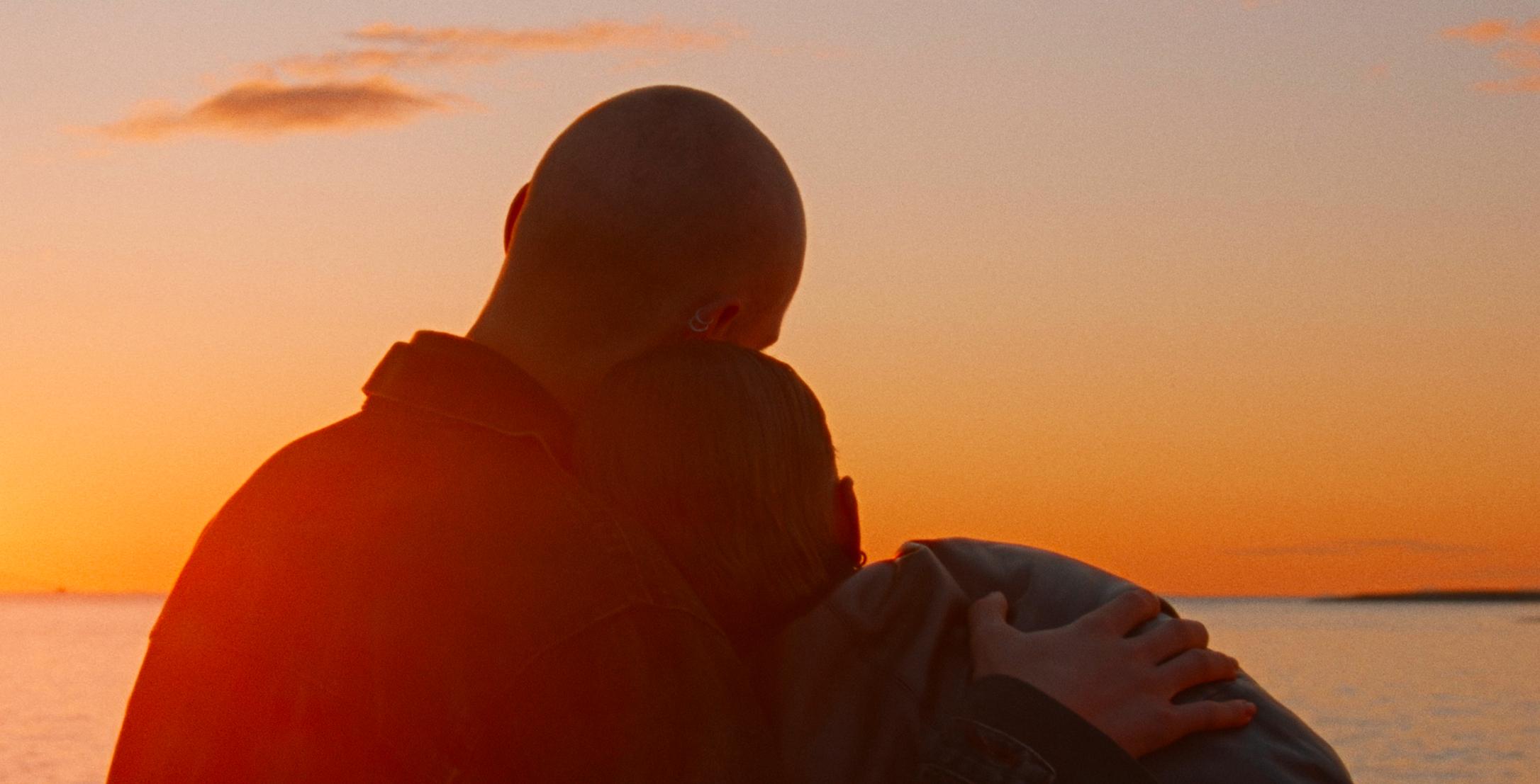 Still aus dem Drama WENN DAS LICHT ZERBRICHT: Im Gegenlicht sitzen ein junger Mann und eine junge Frau am Meer und schauen auf dasLicht der untergehenden Sonne. Der Mann hat den Arm un die Frau gelegt, die sich an ihn anschmiegt. Man sieht beide in der Rückenansicht.