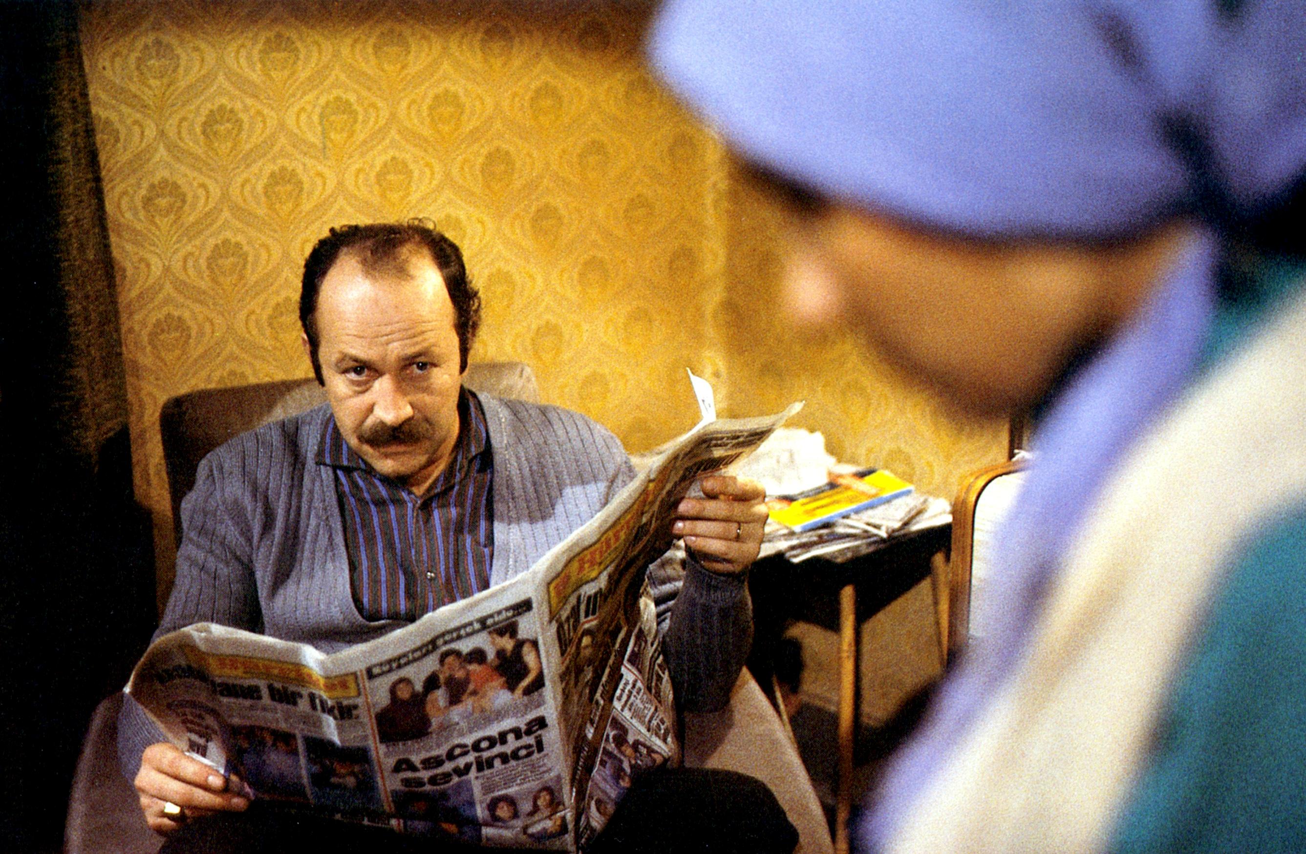 Filmstill aus dem Drama 40qm DEUTSCHLAND: Ein türkischer Mann sitzt in einem Sessel und hält eine Zeitung in der Hand. Er schaut zu einer Frau mit Kopftuch, die im Vordergrund des Bildes unscharf im Profil zu sehen ist.