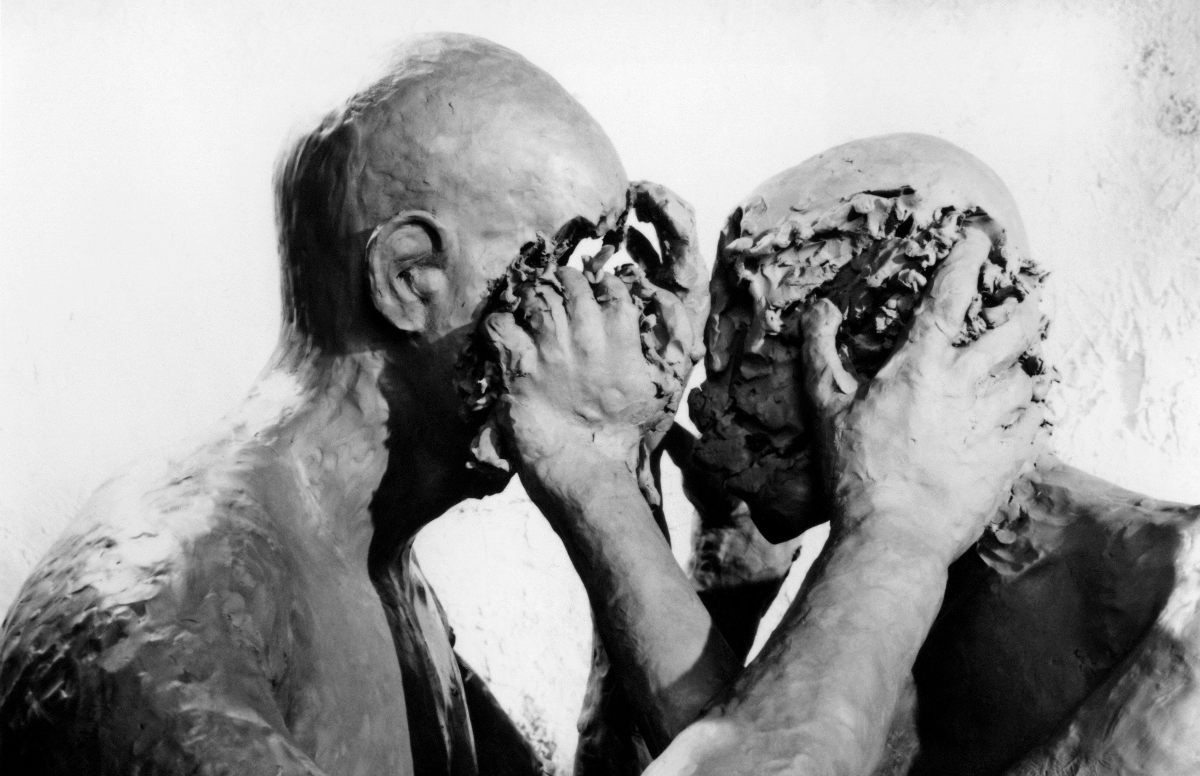 Filmstill aus dem Experimentalfilm TÜCKEN DES GESPRÄCHS: Zwei glatzköpfige Menschen im Profil halten sich an den Köpfen fest. Ihre Gesichter sind verschwunden wirken wie geknetet.