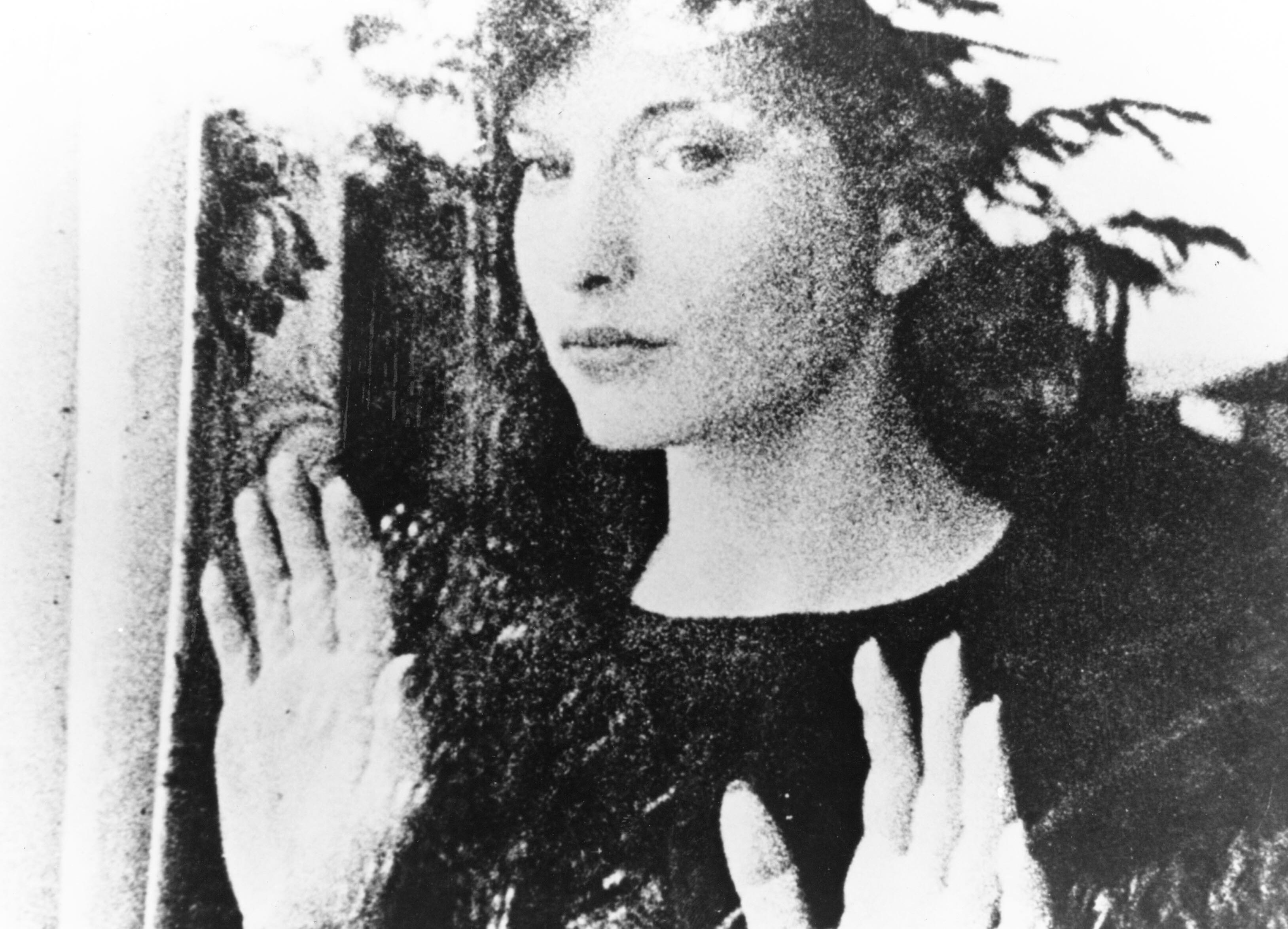 Filmstill aus MESHES OF THE AFTERNOON: Bild einer Frau hinter einer Fesnterscheibe: Sie hält die Hände an das Glas und schaut hinaus. Die Außenwelt spiegelt sich im Glas.