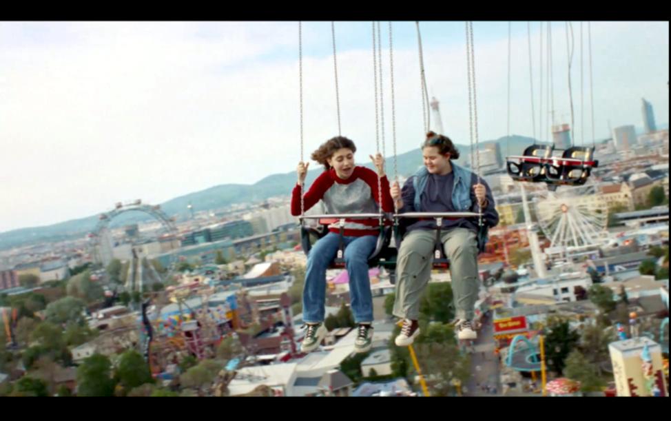 Screenshot aus dem Drama WENN DU ANGST HAST NIMMST DU DEIN HERZ IN DEN MUND UND LÄCHELST: Zwei Mädchen im Teenageralterkreisen in einem Kettenkarussell hoch über einer Stadt.