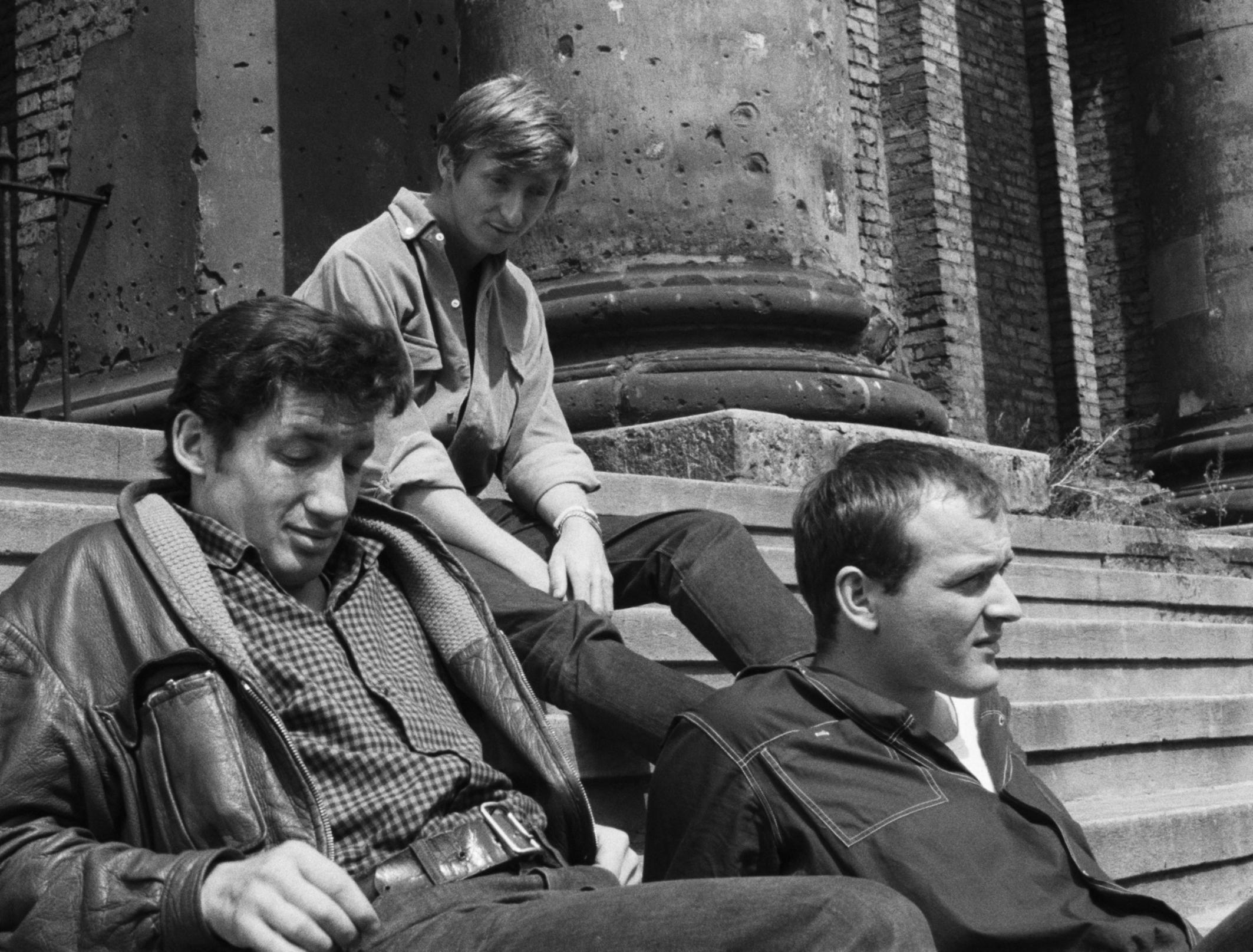 Filmstill aus dem DEFA-Film JAHRGANG 45: Drei junge Männer sitzen auf der Treppe eines Gebäudes, das Einschusslöcher hat.