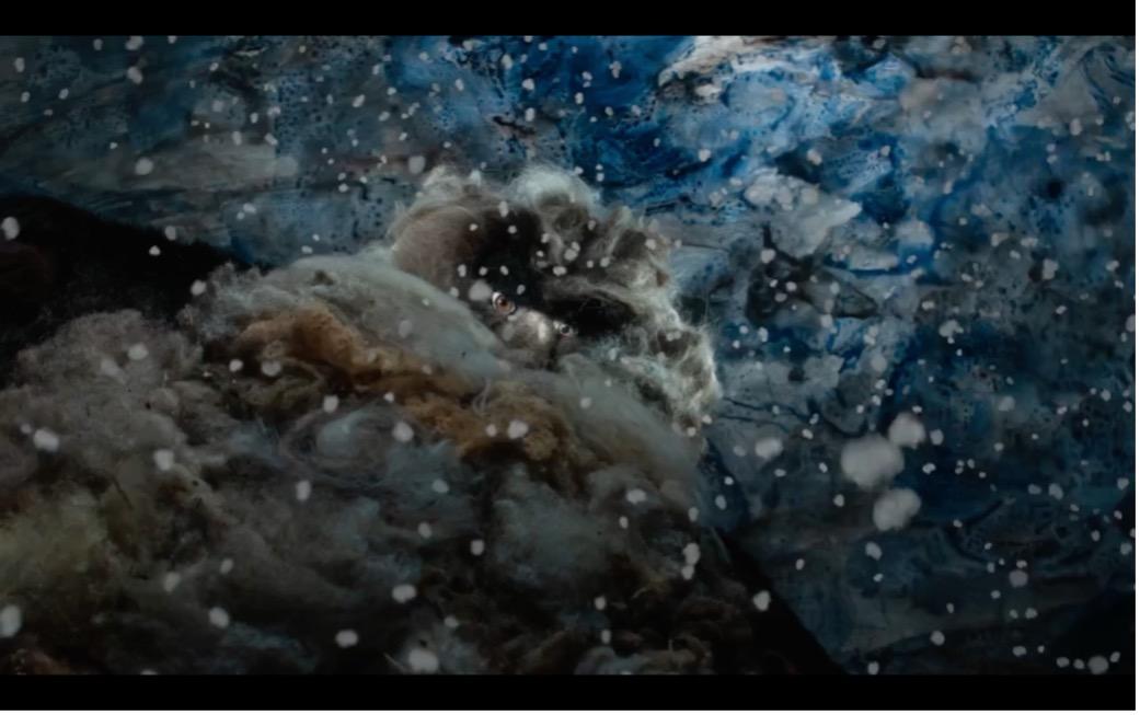 Das Bild zeigt die riesenhafte, trollartige Figur aus größerer Entfernung in einer winterlichen Landschaft. Schneeflocken treiben dicht durch die Szene, während das Wesen fast mit den dunklen Blau- und Grautönen des Hintergrunds verschmilzt. Aus dem dichten Fell und Bart ragen nur die Augen hervor, die aufmerksam und leicht melancholisch wirken.