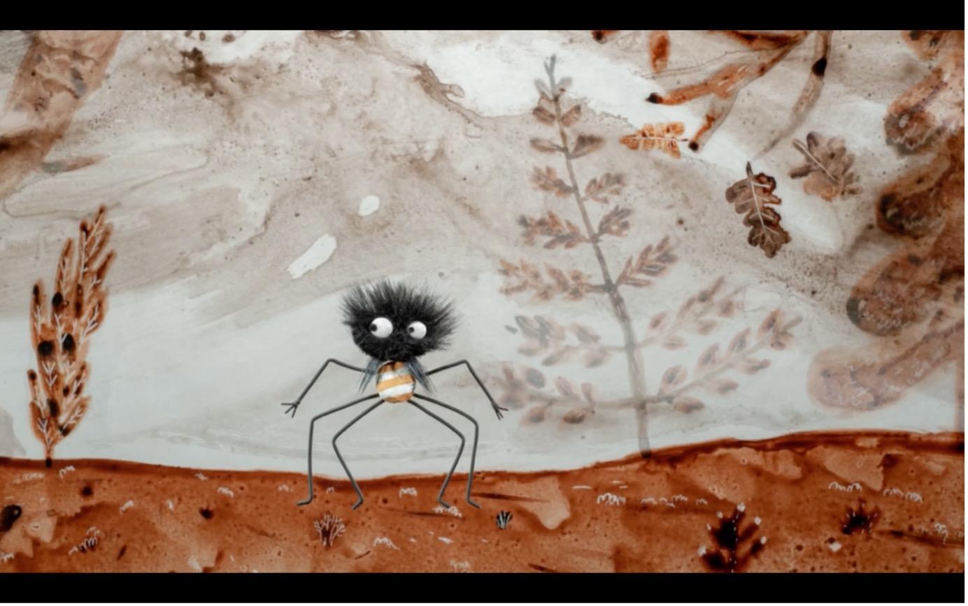 Das Bild zeigt eine stilisierte, fast comicartige Spinnenfigur in einer märchenhaften Landschaft. Die kleine schwarze Spinne besitzt einen kugeligen, zotteligen Körper mit großen weißen Augen und extrem langen, dünnen Beinen. Durch ihren überraschten oder leicht verwirrten Blick wirkt sie eher skurril und liebenswert als bedrohlich.