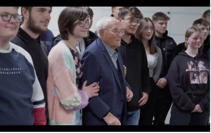 Ein älterer Mann steht inmitten einer Gruppe von Jugendlichen, die sich für ein gemeinsames Foto um ihn versammelt haben. Die körperliche Nähe und die entspannte Haltung der Gruppe vermitteln Vertrautheit und Zugewandtheit.