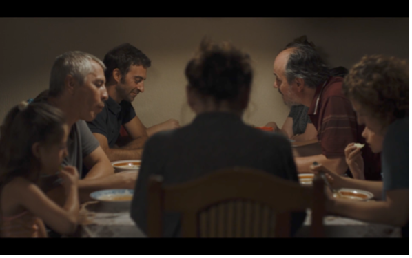 Eine Familie sitzt gemeinsam an einem Tisch und nimmt eine Mahlzeit ein, während sich die Personen einander zuwenden oder auf ihre Teller blicken. Die zentrale Rückenfigur im Vordergrund und die gedämpfte Beleuchtung verleihen der Szene eine ruhige, zugleich leicht angespannte Atmosphäre.