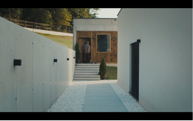 Ein Mann steht am Eingang eines modernen Hauses, zu dem ein langer, geradliniger Weg zwischen hohen Betonwänden führt. Die strenge, symmetrische Bildkomposition und die dominante Architektur lassen die einzelne Figur klein und beinahe verloren erscheinen.