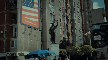 Vor einem hohen Wohngebäude steht eine Statue Bill Clintons, daneben hängt ein großformatiges Plakat mit der US-Flagge und der Aufschrift „Faleminderit Bill Clinton“. Die amerikanischen Symbole prägen das Stadtbild und verweisen auf die besondere Bedeutung der USA für die jüngere Geschichte des Kosovo.