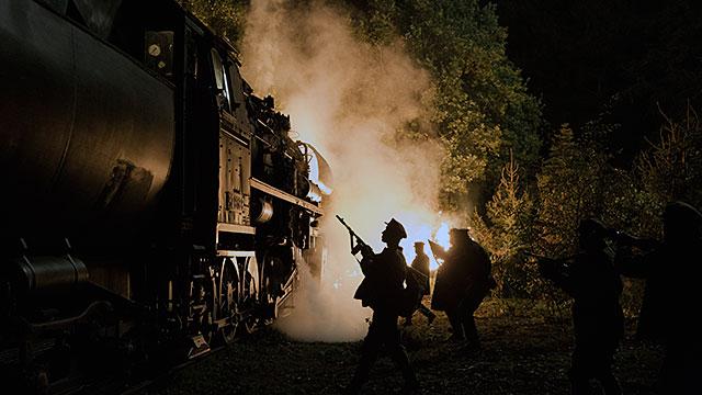Babylon Berlin, Szenenbild: Zugüberfall in der Nacht, der weiße Dampf der Lok hebt sich vom dunklen Hintergrund ab. Im Vordergrund steht schemenhaft ein bewaffneter Mann